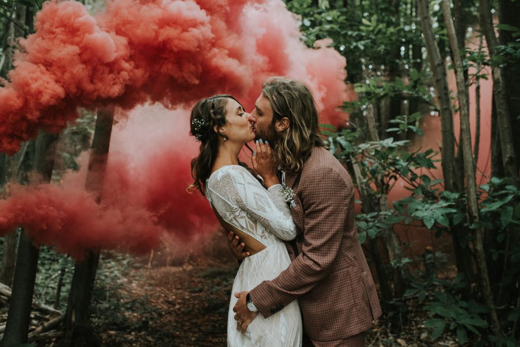 Image of a couple kissing in a forest surrounded by red smoke taken by Fern Edwards